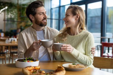 Sıcak bir restoranda kahve ve yemek paylaşan çift ahşap masada salata ve pizza tabağıyla oturuyor.