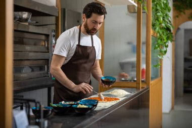 Pizza sosu. Erkek fırıncı pizzacı mutfağında çalışıyor ve pizzaya malzeme koyuyor.