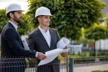 İnşaatçılar bina incelemesi sırasında ağaçlı ve doğal çevresi olan açık yeşil bir alanda planlar ve dizüstü bilgisayarları tartışıyorlar.