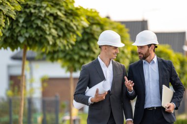 Açık havada yeşillik ve doğal ışıkla bezenmiş verimli bir ortamda taslak ve dizüstü bilgisayarları inceleyen sert şapkalar takan mühendisler.
