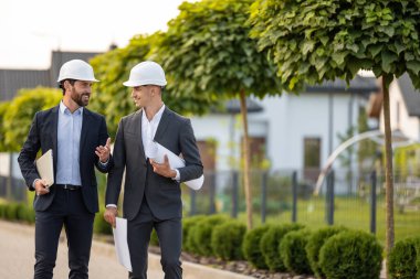 İnşaat ekibi proje planlaması sırasında yeşil alanların ortasındaki bina planlarını ve dizüstü bilgisayarlarını analiz ediyor.