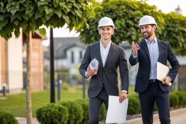 Açık havada yeşillik ve doğal ışıkla bezenmiş verimli bir ortamda taslak ve dizüstü bilgisayarları inceleyen sert şapkalar takan mühendisler.