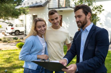 Emlakçı, banliyödeki konutların önünde çiftlere ait belgelerle birlikte pano sunuyor.