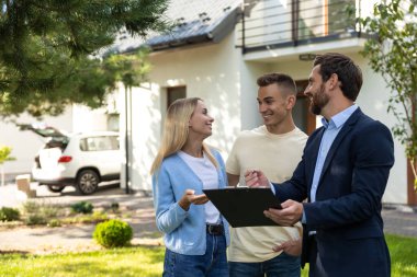 Emlakçı, konut alanının önünde pano kullanan bir çiftle emlak detaylarını tartışıyor.