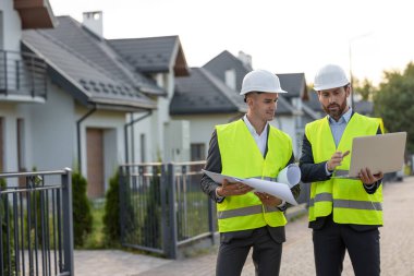 Kasketli ve güvenlik yelekli inşaat işçileri banliyödeki modern konutların önündeki planlar ve dizüstü bilgisayarları inceliyorlar.