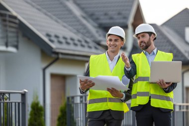 Güvenlik yelekleri ve başlık mühendisleri modern konut binalarının önündeki projeleri planlar ve dizüstü bilgisayarlarla koordine ediyorlar.