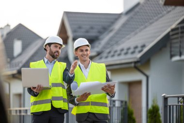 İnşaat ekibi inşaat başlıkları ve yelekleriyle yerleşim bölgesindeki modern evlerin önünde ayrıntılı planlar ve dizüstü bilgisayarlarla bölgeyi inceliyor.