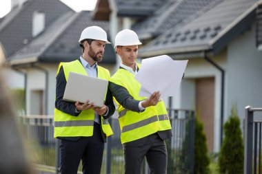 Kasketli ve güvenlik yelekli inşaat işçileri banliyödeki modern konutların önündeki planlar ve dizüstü bilgisayarları inceliyorlar.
