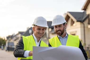 Kasketli ve güvenlik yelekli inşaat işçileri banliyödeki modern konutların planlarını gözden geçiriyorlar.