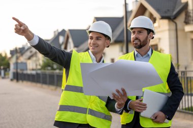 Kasket ve güvenlik yelekleri takan inşaatçılar, banliyödeki evlerin önündeki planlar ve dizüstü bilgisayarları analiz ediyorlar.