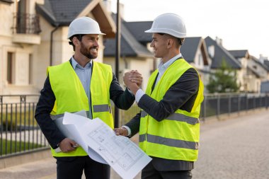 Kasket ve güvenlik yelekleri takan inşaatçılar banliyödeki evlerin önündeki planları analiz ediyorlar.