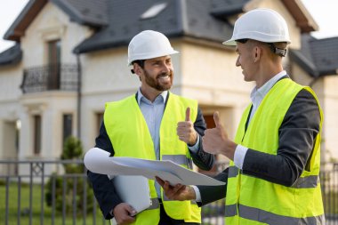 Kasketli ve güvenlik yelekli inşaat işçileri banliyödeki modern konutların planlarını inceliyorlar.