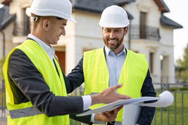 Kasket ve güvenlik yelekleri takan inşaatçılar banliyödeki evlerin önündeki planları analiz ediyorlar.