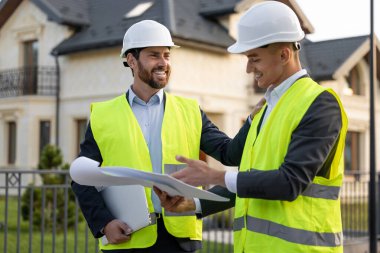 Kasket ve güvenlik yelekleri takan inşaatçılar banliyödeki evlerin önündeki planları analiz ediyorlar.