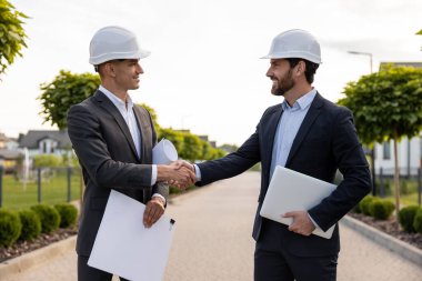 Sert şapkalar takan mühendisler, yeşil çevre koşulları olan evlerin önünde planlar tutarken el sıkışarak anlaşmayı mühürlüyorlar.