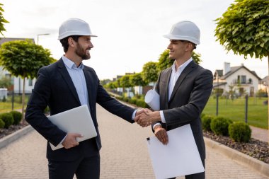 Mühendisler, açık hava ortamında yemyeşil evlerin ortasında bina planları ve dizüstü bilgisayarlarıyla el sıkışıyorlar.