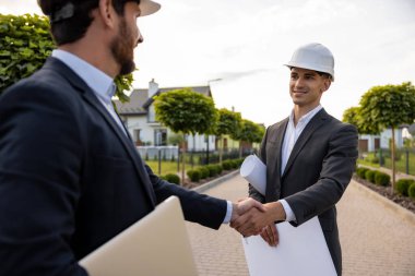 Sert şapkalar takan mühendisler, yeşil çevre koşulları olan evlerin önünde planlar tutarken el sıkışarak anlaşmayı mühürlüyorlar.