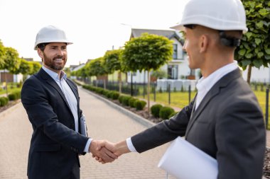 İnşaat ekibi inşaat başlıklarıyla projeyi el sıkışarak kutlarken aynı zamanda yeşil çevresi olan evlerin yakınındaki planları tutuyor.