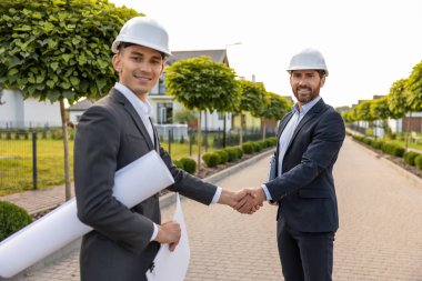 Mühendisler, açık hava ortamında yemyeşil evlerin arasında bina planlarıyla el sıkışıyorlar.