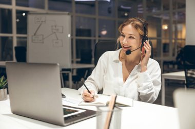 Ofis çalışanı, yeşil kulaklıklı parlak odada dizüstü bilgisayardaki görevleri yönetiyor.