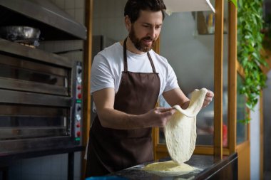 Beyaz tişörtlü ve kahverengi önlüklü sakallı bir adam fırın mutfağında un tezgahı ve rafta bitkilerle pizza hamuru atıyor.