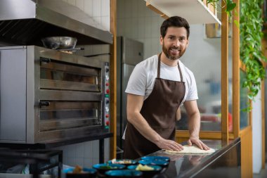 Beyaz gömlekli, siyah önlüklü, sakallı bir adam mutfakta pizza için hamur esnetiyordu.