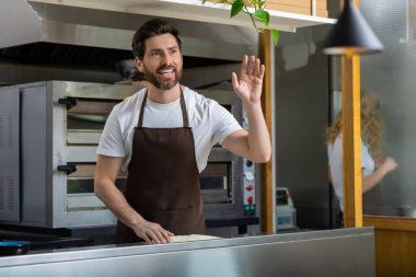Önlüklü, beyaz tişörtlü, gülümseyen, sakallı, fırıncı bir erkek.