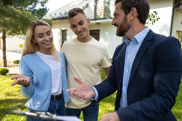 Emlakçı, konut alanının önünde pano kullanan bir çiftle emlak detaylarını tartışıyor.