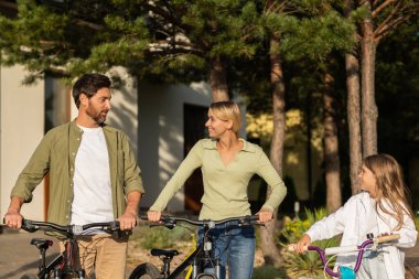 Güneşli bir günde modern bir garajın önünde bisiklete binmeye hazırlanan bir aile.