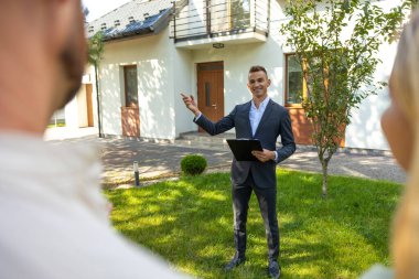 Emlakçı, güneşli bahçede çiftlere modern ev ve yeşillik sunuyor.