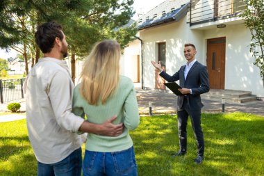 Emlakçı, güneşli bahçede çiftlere modern ev ve yeşillik sunuyor.