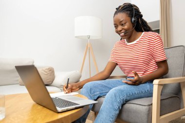 Çevrimiçi çalışma. Kulaklıklı genç bir kadın dizüstü bilgisayarda oturuyor ve meşgul görünüyor.