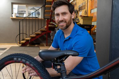 Motosiklet tamircisi atölye girişinde duruyor. Üzerinde mavi tişört ve hız ustası bisiklet ekipmanları var.