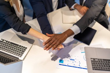 Toplantı masasında işbirliği yapan çok ırklı profesyoneller grubu proje üstüne olumlu bir jest yaparak el ele tutuşuyor.