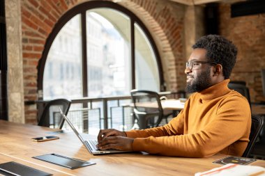 Gözlüklü sakallı adam dizüstü bilgisayarda yazıyor. Endüstriyel tarzda şık bir ofiste çalışıyor.