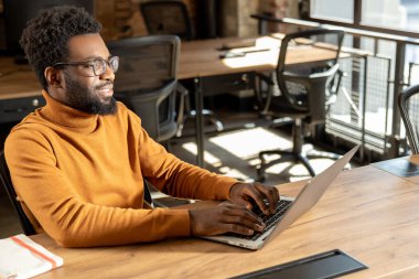 Gözlüklü sakallı adam dizüstü bilgisayarda yazıyor. Endüstriyel tarzda şık bir ofiste çalışıyor.