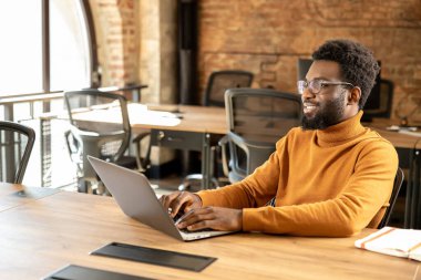 Uzaktan kumandalı bilgisayarla çalışan Afro-Amerikan erkek profesyonel. Kemerli pencereleri ve tuğlalı duvarları olan parlak bir çalışma alanında çalışıyor.