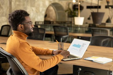 Turuncu balıkçı yaka kazaklı erkek serbest işçi modern çalışma alanındaki ahşap masada oturan projeye odaklanmış durumda.