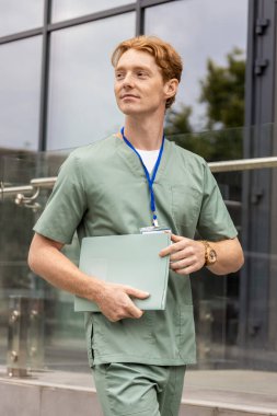 Laboratuvar önlüklü kızıl saçlı sağlık çalışanı hasta geçmişi olan cam binanın önünden geçiyor.