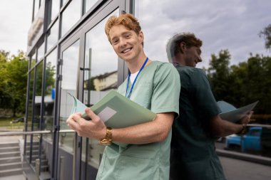 Cam klinik binasının yanında dikilen laboratuvar önlüklü kızıl doktor elinde klinik geçmişi olan bir dosya tutuyor.