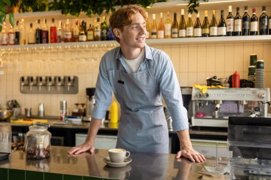 Bar tezgahına hafifçe yaslanmış, hoş bir kahve dükkanının içinde dişleriyle gülümseyerek duran memnun barista.