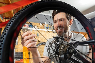 Profesyonel tamir atölyesinde bisiklet tekerleği tutan gülümseyen erkek teknisyen. Çekmeceler ve metal merdivenlerle çevrili.