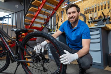 Mavi polo tişörtlü, gülümseyen sakallı adam arka planda kırmızı merdiven ve aletlerle modern kapalı atölyede bisiklet tekerleğini tamir ediyor.