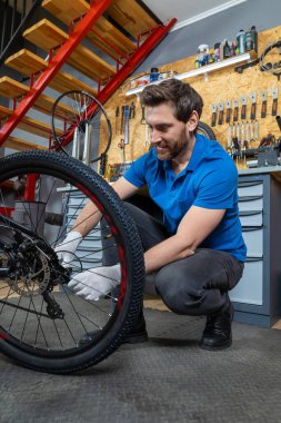 Sakallı adam bisiklet bakım sırasında çekmeceler ve endüstriyel merdivenlerle çevrili kapalı atölyedeki bisiklet tekerine servis yapıyor.