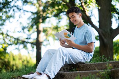 Parkta kitap okuyordum. Koyu saçlı pozitif genç parkta merdivenlerde oturuyor.