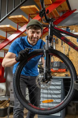 Mavi polo takımlı sakallı adam atölyedeki bisiklet tekerleği raflar ve tamir ekipmanlarıyla çevrili.