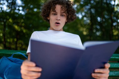 Şaşırdım. Kıvırcık saçlı genç parkta bankta oturuyor ve kitap okuyor ve şaşırmış görünüyor.