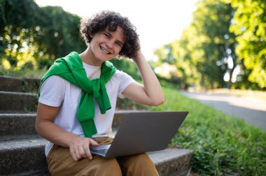 Görüntülü arama. Kıvırcık saçlı genç parkta merdivenlerde oturuyor ve bir video görüşmesi yapıyor.
