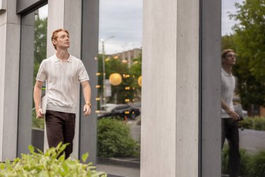 Kızıl saçlı, yirmili yaşlarda bir elinde dizüstü bilgisayar taşıyan bir adam binanın etrafında hareket ediyor.