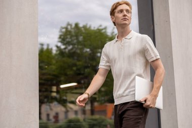 Yirmili yaşlarda genç bir erkek elinde modern ofis binasının yanındaki bir dizüstü bilgisayarla sokakta yürüyor.